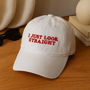 以下が含まれることがあります： 赤い文字で「I JUST LOOK STRAIGHT」と刺繍された白い野球帽。 つばが湾曲しており、クラシックな6パネルデザインです。 帽子は木製の表面に置かれています。