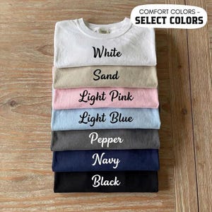 May include: A stack of seven folded t-shirts in various colors including white, sand, light pink, light blue, pepper, navy, and black. Each shirt features its color name printed in black script text. A label at the top reads COMFORT COLORS - SELECT COLORS.