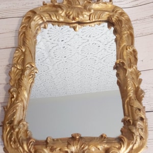 May include: A rectangular gold-colored mirror with ornate, scrolling details. The frame has a baroque style with a decorative top and bottom. The mirror reflects a white lace pattern and a plain wall. The mirror is likely for home decor.