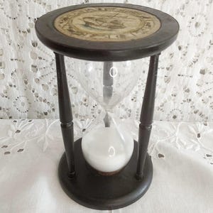 Puede incluir: Un reloj de arena antiguo con un marco de madera oscura y un centro de vidrio transparente. La parte superior e inferior son circulares, y el reloj de arena está sostenido por cuatro patas delgadas. La parte superior presenta una ilustración descolorida. Arena blanca fluyendo.