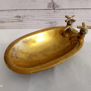 May include: Oval-shaped brass dish with two faucet-shaped handles. The dish has a hammered texture and a golden hue. The faucets are detailed with cross-shaped handles. The background is a light-colored wooden surface.