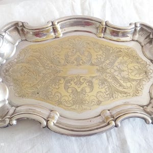May include: An oval silver-toned serving tray with a scalloped, raised edge and a central gold-toned inlay featuring intricate, symmetrical engraved scrollwork and floral patterns across the flat surface of the metal serving piece.