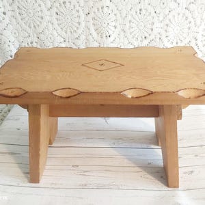 Op de afbeelding: Een kleine, lichtgekleurde houten kruk met een rechthoekige bovenkant en vier poten. De bovenkant heeft een decoratieve rand met gesneden details en een ruitvormige inleg. De kruk is ongeveer 30 cm lang, 20 cm breed en 15 cm hoog.