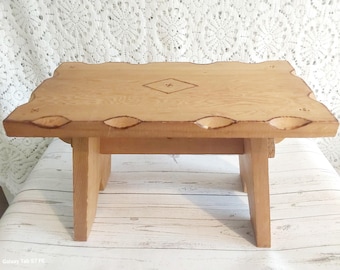 May include: A small, light-colored wooden step stool with a rectangular top and four legs. The top has a decorative edge with carved details and a diamond-shaped inlay. The stool is approximately 12 inches long, 8 inches wide, and 6 inches tall.