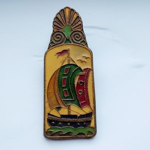 Könnte beinhalten: Eine rechteckige Vintage-Brosche mit Segelboot-Design. Die Brosche hat einen gelben Hintergrund mit einem Schiff mit grünen, roten und goldenen Segeln. Die Oberseite der Brosche hat ein Sonnenstrahl-Design. Die Brosche ist wahrscheinlich aus Metall.