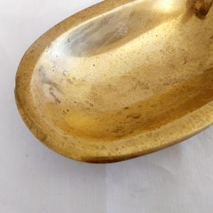 May include: A close-up shot of a vintage, oval-shaped brass dish. The dish has a tarnished, aged appearance with a golden-brown hue. The edges are slightly raised, and the interior shows signs of wear.