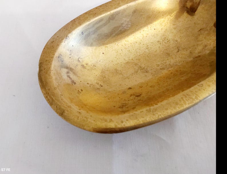 May include: A close-up shot of a vintage, oval-shaped brass dish. The dish has a tarnished, aged appearance with a golden-brown hue. The edges are slightly raised, and the interior shows signs of wear.