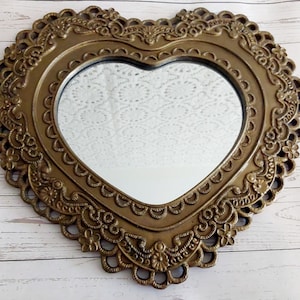 May include: A heart-shaped mirror with an ornate bronze frame. The frame is decorated with detailed floral and scroll patterns. The mirror has a smooth, reflective surface. The design is vintage in style.