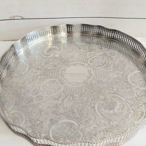 Könnte beinhalten: Ein rundes Serviertablett aus silberfarbenem Metall mit einem detaillierten, geprägten Blumen- und Rankenmuster auf der Oberfläche. Das Tablett hat einen erhöhten, gewellten Rand mit einem vertikalen Schlitzdesign am gesamten Umfang.