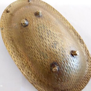 May include: A vintage, oval-shaped brass dish with a textured surface and four small, bolted feet. The dish has a hammered appearance, with a warm, golden hue. The design suggests a decorative or functional piece, possibly for display or serving.