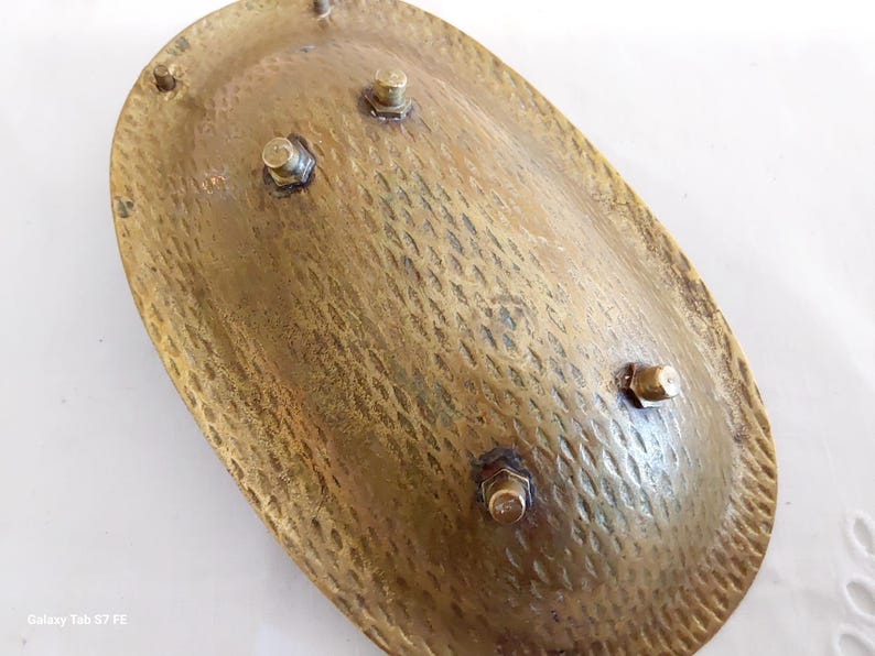 May include: A vintage, oval-shaped brass dish with a textured surface and four small, bolted feet. The dish has a hammered appearance, with a warm, golden hue. The design suggests a decorative or functional piece, possibly for display or serving.