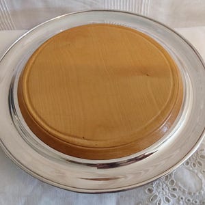 May include: A silver serving tray with a light brown wooden insert. The tray has a wide, reflective rim and a circular wooden centre. The wood has a natural grain pattern.