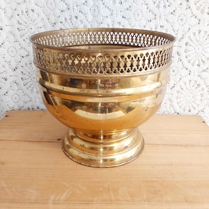 May include: A vintage brass planter with a decorative openwork rim. The planter has a rounded bowl shape and sits on a pedestal base. The brass has a warm, golden hue, and is likely used for displaying plants or flowers.