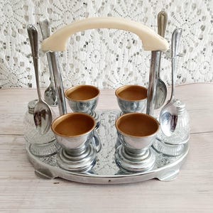 Può includere: Un set di portauova vintage argentato con quattro portauova, cucchiai e saliere. Il set include una maniglia color crema ed è presentato su un vassoio a specchio. I portauova hanno un esterno argentato e un interno dorato.