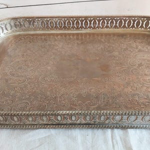 Könnte beinhalten: Ein rechteckiges Tablett aus silberfarbenem Metall mit einem detaillierten, geprägten Blumen- und Rankenmuster auf der Basis. Das Tablett hat einen erhöhten, durchbrochenen Rand mit ovalem Muster und eine texturierte Perlenkante.