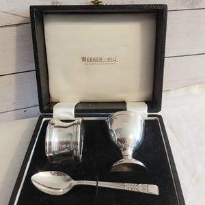 Pode incluir: Um porta-ovos, um anel de guardanapo e uma colher de metal prateado apresentados numa caixa preta com dobradiças. O interior da tampa apresenta o texto Webber and Hill Cutlers and Silversmiths. Os itens estão sobre uma base de veludo preto.