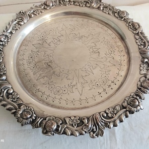Peut inclure: Un plateau de service rond en métal argenté doté d'une bordure ornée de motifs floraux et de feuilles en relief. Le centre du plateau présente un motif gravé complexe composé de feuilles stylisées et de petites fleurs en cercle.