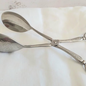 May include: A pair of silver-plated serving tongs featuring two spoon-shaped ends, one of which has a decorative floral engraving. The handles are long and slender with an ornate pattern, ending in two circular finger loops for gripping.