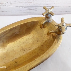 May include: A vintage brass bathtub-shaped dish with two cross-shaped faucet handles. The dish has an oval shape and a tarnished, aged appearance, suggesting it is an antique decorative item.