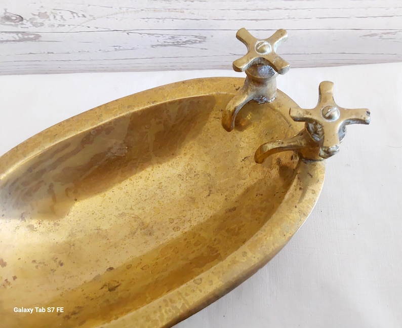 May include: A vintage brass bathtub-shaped dish with two cross-shaped faucet handles. The dish has an oval shape and a tarnished, aged appearance, suggesting it is an antique decorative item.