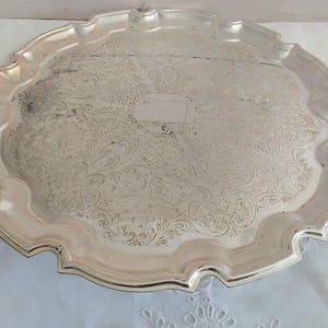 May include: A silver-coloured, round serving tray with a decorative, scalloped edge. The tray features an ornate, embossed floral pattern across its surface. The centre of the tray has a rectangular area. The tray is displayed on a white surface.