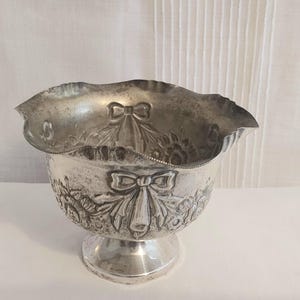 Puede incluir: Un cuenco plateado con borde festoneado y base de pedestal. El cuenco presenta diseños en relieve de flores y lazos en el exterior. El cuenco es probablemente de metal y es adecuado para fines decorativos.