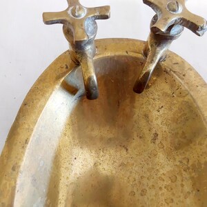 May include: A vintage brass soap dish with two faucet-style handles. The dish has an oval shape and a tarnished, aged appearance. The faucets are cross-shaped and add a decorative touch to the piece.