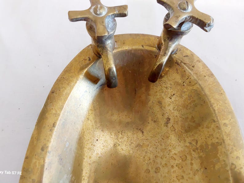 May include: A vintage brass soap dish with two faucet-style handles. The dish has an oval shape and a tarnished, aged appearance. The faucets are cross-shaped and add a decorative touch to the piece.