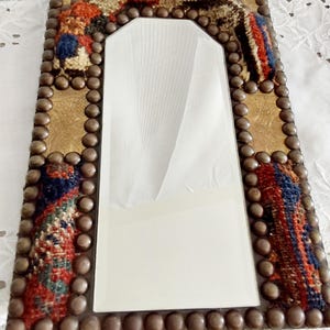 May include: A rectangular mirror with a decorative frame. The frame is made of wood and is covered in a colourful fabric with a geometric pattern. The frame is also decorated with metal studs.