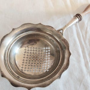 May include: A silver-toned metal strainer with a scalloped edge and a central square section featuring a grid of small cross-shaped perforations. The strainer includes a long, slender metal handle topped with a smooth, light brown wooden grip.