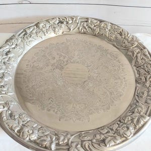 May include: A round silver-toned metal serving tray featuring an intricate embossed floral border with leaves and blossoms. The centre of the tray displays a detailed engraved scrollwork pattern with a smooth, circular area in the middle.