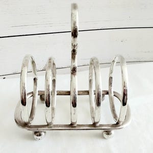 May include: A silver-toned metal toast rack featuring four vertical rounded slots for holding slices of bread. The rack has a central arched handle for carrying and is supported by four small spherical feet at the base of the rectangular frame.