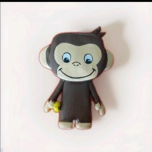 Könnte beinhalten: Eine braune Affenfigur mit lächelndem Gesicht, blauen Augen und beigefarbenen Ohren. Der Affe hält eine gelbe Banane. Die Figur steht auf einer hellrosa Oberfläche.