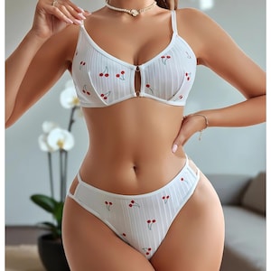May include: White lingerie set with a cherry pattern. The set includes a bra with adjustable straps and a matching thong. The bra has a keyhole detail in the center. The fabric has a ribbed texture.