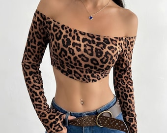 May include: An off-the-shoulder long-sleeve crop top featuring a brown and black leopard print pattern. The garment is paired with blue denim jeans and a brown leather belt with a gold-toned buckle. The word BRAVURE is printed in the top left corner.