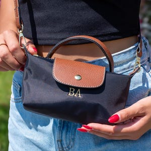 May include: A black crossbody bag with a brown leather flap and handle. The bag has the initials "BA" in gold. It has a gold chain strap. The bag is held by a person wearing a black top and blue jeans.