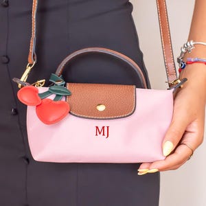 Puede incluir: Un bolso rosa con solapa y asa marrones, con las iniciales "MJ" en rojo. El bolso tiene una correa marrón y está adornado con un charm de cereza. La persona lleva un vestido negro y pulseras doradas.
