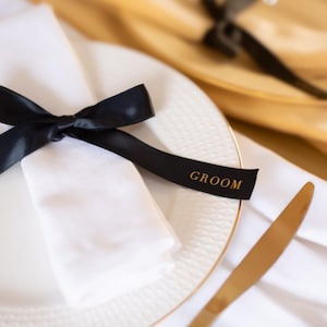 Puede incluir: Decoración de mesa formal para una boda. Plato blanco con borde dorado, servilleta blanca y cinta negra con la palabra "GROOM" en dorado. Un cuchillo dorado a la derecha.