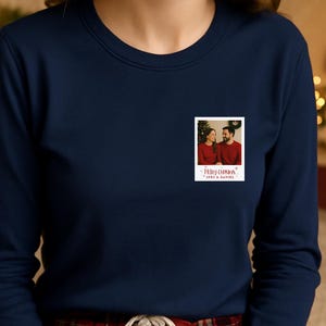 May include: Navy blue sweatshirt with a small, rectangular photo print. The photo shows a couple in red shirts with a Christmas tree in the background. The text "Merry Christmas" and names are below the photo.