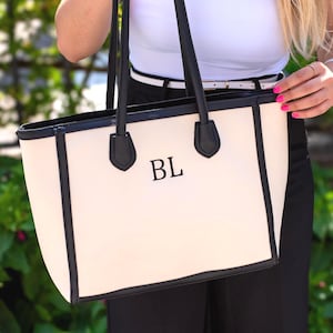May include: Cream tote bag with black trim and handles. The bag has the initials "BL" in black. The handles are attached with black leather accents. The bag is held against a background of green foliage.