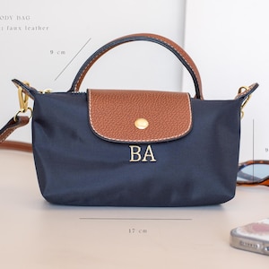 May include: Navy blue crossbody bag, 17 cm wide and 9 cm high, with a brown flap and handle. The bag features the initials "BA" in gold lettering. The material is faux leather.