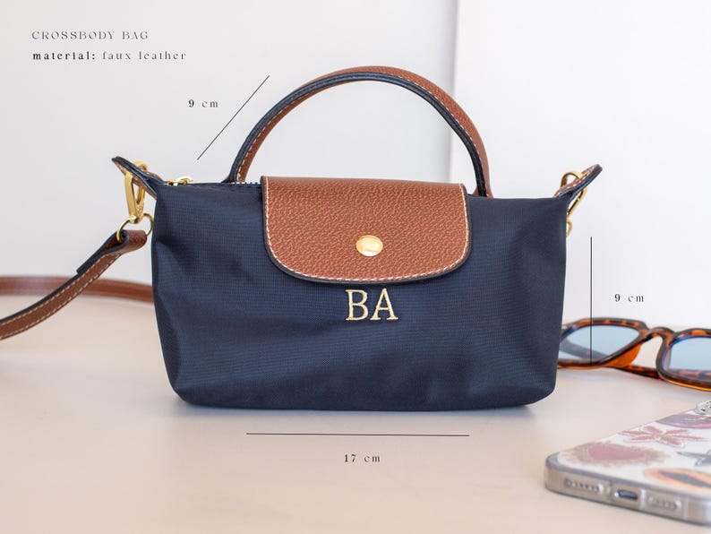 May include: Navy blue crossbody bag, 17 cm wide and 9 cm high, with a brown flap and handle. The bag features the initials "BA" in gold lettering. The material is faux leather.