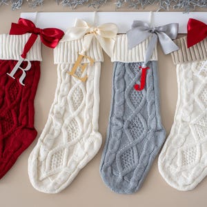 Peut inclure: Quatre bas de Noël en tricot torsadé, rouges, blancs et gris, chacun avec une lettre différente. Les bas sont accrochés à un support en bois blanc et ornés de nœuds en satin rouges, crème et gris.
