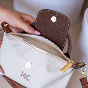 May include: Cream-colored crossbody bag with brown leather accents and a gold zipper. The bag features a flap closure and a shoulder strap. The initials "MC" are embroidered on the front. The bag is open, revealing the interior.
