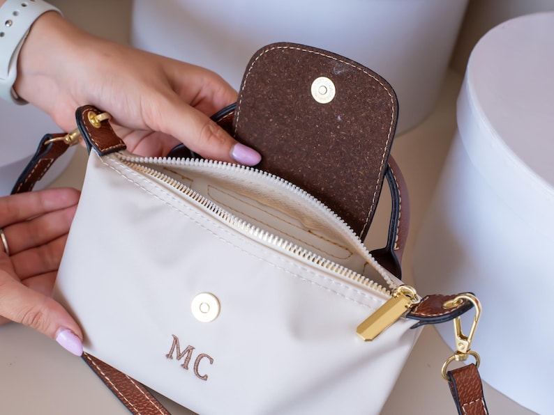 May include: Cream-colored crossbody bag with brown leather accents and a gold zipper. The bag features a flap closure and a shoulder strap. The initials "MC" are embroidered on the front. The bag is open, revealing the interior.