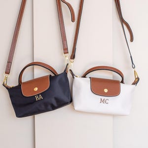 May include: Two small shoulder bags, one navy blue and one white, each with a brown flap and handle. The navy bag has "BA" embroidered, the white bag has "MC". Both have brown straps and gold hardware.
