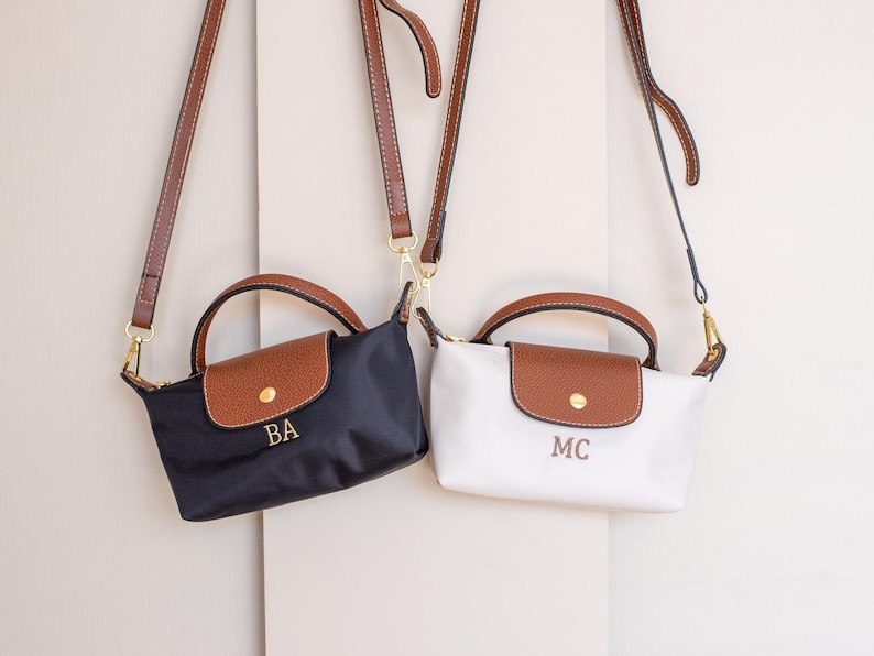 May include: Two small shoulder bags, one navy blue and one white, each with a brown flap and handle. The navy bag has "BA" embroidered, the white bag has "MC". Both have brown straps and gold hardware.