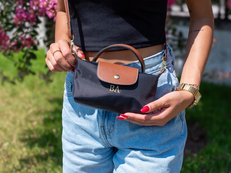May include: Black crossbody bag with a brown flap and handle. The bag features the initials "BA" in gold lettering. The bag has a gold chain strap and a gold button closure. The bag is held by a person wearing a black top and blue jeans.