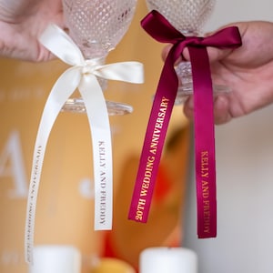 May include: Two champagne flutes with decorative ribbons. One ribbon is white, the other is burgundy. Both ribbons have gold text that reads "KELLY AND FREDDY" and "20TH WEDDING ANNIVERSARY". The flutes are held up in a celebratory toast.