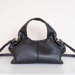 Könnte beinhalten: Schwarze Lederhandtasche mit runder Form und oberem Griff. Die Tasche hat eine strukturierte Oberfläche, einen langen, verstellbaren Riemen und goldfarbene Beschläge. Die Tasche steht auf einer weißen Oberfläche.